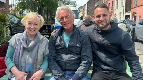 Three people sit on a picnic table beside a busy city street. A woman is on the left, wearing a scarf and green jumper, the man in the middle has longish blond hair and is weraing jeans and a blue jacket, while the man on the right has short brown hair and a short beard. He is wearing a blue jacket