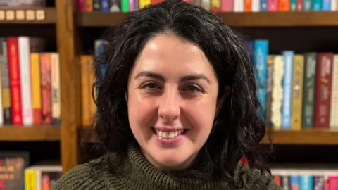 A woman - Zoe Biles - smiling straight at the camera. She is wearing a khaki jumper and standing in front of full bookshelves.