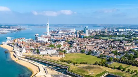 Getty Images Aerial view of the historic Long Curtain Battery coastline in Portsmouth, UK. The city is a mix of low-rise housing and a few tower blocks in the centre. It is on the edge of Portsmouth Harbour and the white Spinnaker Tower is visible in the centre of the picture. The sky is blue and it is a sunny day.