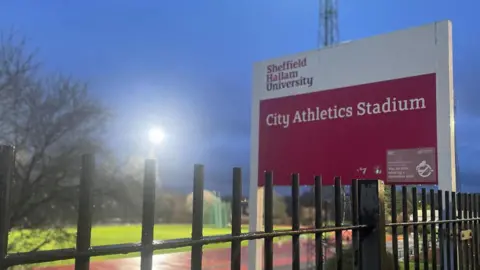 A large metal black fence. Behind it is a wet athletics track slick with rain lit by floodlights