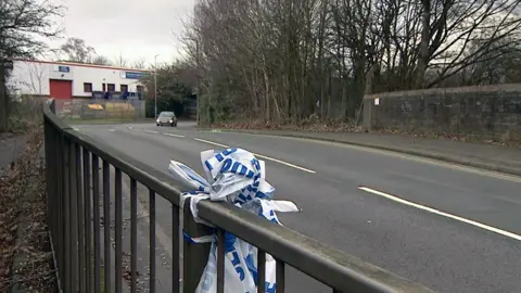 BBC Police tape tied on to a metal bridge on a curved industrial road lined with trees and bushes. There is white warehouse in the background.
