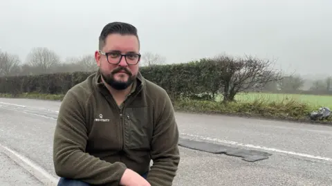 A man crouching on a pavement with a repaired pothole on the road in the background