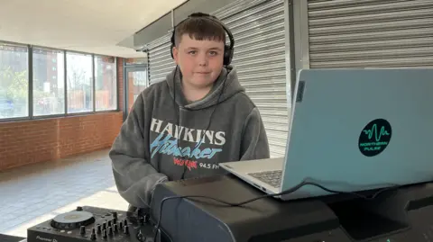 Kay Davidson/BBC Albie is wearing headphones and a grey sweatshirt with 'Hawkins Hitmaker' written on the front. He is standing behind a silver laptop and DJ decks and in front of a window and a closed shop front. 