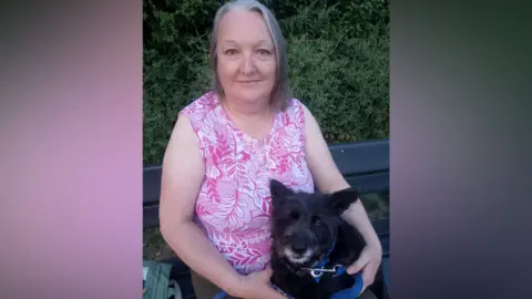 Supplied A woman with shoulder length grey hair is wearing a pink and white sleeveless blouse with a floral pattern as she sits on a black bench with her small black dog on her lap, with a green hedgerow seen behind her.