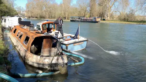 BBC A man stood on an active boat parked next to a burnt out canal boat. There is piping running into the abandoned boat as it is prepared for removal.