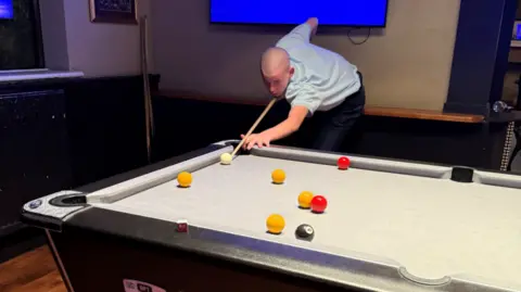 Family A boy with a bald head, light blue shirt and black trousers leans over a pool table while pointing a cue at a ball. 