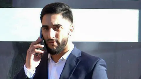 Spindrift Akbar Razaq, who has black hair and a black beard, is holding a mobile phone t his ear as he looks at the camera. He is wearing a dark blue jacket and white shirt