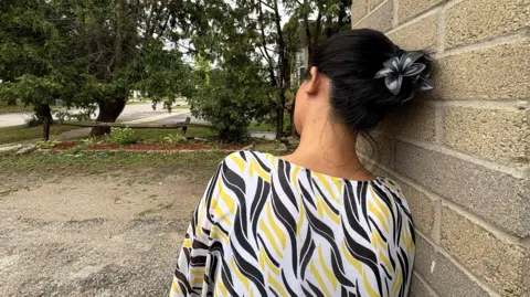 BBC News A photo of the woman interviewed in the story. It does not show her face and is taken from behind. Her hair is black and is held up by a black clip shaped like a flower. She is wearing a blouse with yellow, black and white print. She leans on a brick wall, and is overlooking a suburban road lined by trees.