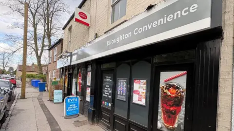 Kevin Shoesmith/BBC A black-fronted village shop, with cars park outside. The signs says, 'Elloughton Convenience'. In the foreground, the shop front features a large photograph of a glass of coke.