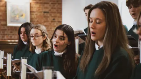 Singers from the University of Exeter's chapel choir performing a song. Four young women are in the front row wearing green tunics. A couple of male singers wearing the same clothes are behind them.