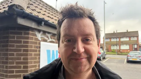 A man stands outdoors beside a small brick building with a tiled roof and graffiti on the wall. Cars and rows of red-brick houses are visible in the background under an overcast sky. The person is wearing a dark jacket over layered clothing. He has brown hair.
