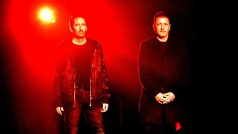 Getty Images Nine Inch Nails members Trent Reznor and Atticus Ross stand against a dark background, illuminated by a red light.