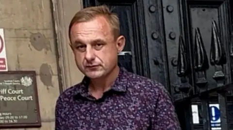 A man in his early 40s outside a court building. He has short blonde hair and is wearing a patterned purple shirt