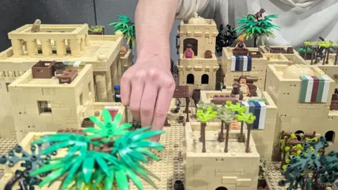 Christian Resources Exhibition Mainly cream-coloured miniature model of ancient stone-built buildings with palm trees and a few people.  A hand is reaching down onto the model.