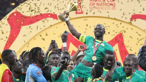 Sadio Mane, held aloft by several Senegal team-mates, holds the golden Africa Cup of Nations trophy aloft in his right arm. Mane wears an all-green Senegal kit, a yellow captain's armband on his right bicep and a gold medal on a red ribbon around his neck. Gold ticker tape falls in the background in front of a yellow and red background as Senegalese celebrate around Mane