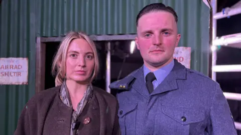 Leah and Glenn are in costume. Leah has blond hair and is wearing a brown coat. Glenn has a buzz cut and is dressed in an RAF uniform. They're standing on stage, with a notice on a metal wall saying air raid shelter.