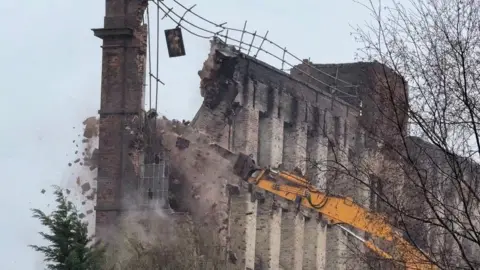 The wall of a historic mill building is being pulled down by a yellow mechanical device. There are bricks and debris falling as the wall begins to collapse.