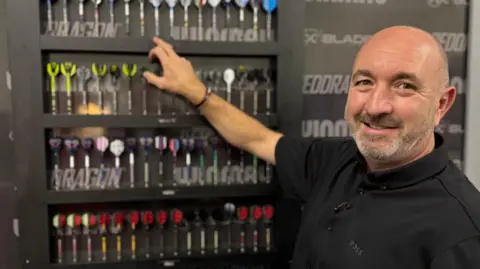 Head of development Lee Huxtable, is pictured next to cabinet which contains five rows of darts. He is pointing at one dart, and is wearing a black polo shirt. Lee is bald with short stubble.