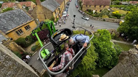 Aerial view of two experts reinstalling the clock at St Catherine's Church, in Montacute, Somerset. A crowd looks on from the street below.