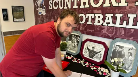 Jamie Benson signs the book of condolence which is placed on a table in front of three black and white pictures of Keegan in frames with flowers in front. Benson has short black hair and beard and is wearing a red shirt.