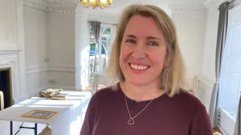 Nailsea historian Dr Jo Edwards, who has blonde, shoulder-length hair and is wearing a burgundy jumper and a gold heart necklace. She is smiling at the camera. She is in a white room with bay windows and a desk behind her with artefacts on the table. 