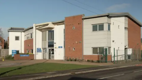 BBC/Ed Young A modern, two-story building with a combination of red brick and white panel exterior walls. The structure has a contemporary design featuring clean lines and a slightly curved roof section on the left side. Large windows are visible on both floors, allowing natural light into the building. The main entrance is located at the centre, marked by a glass door and flanked by blue signage. There is a paved pathway leading to the entrance, bordered by a small landscaped area with greenery. Surrounding the building is a metal fence, and the foreground includes a road with a pedestrian crossing. In the background, there are residential houses and leafless trees.