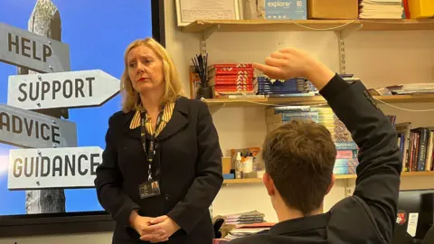 Gillian stands at the front of a classroom near a screen displaying the words “Help,” “Support,” “Advice,” and “Guidance,” while students sit at desks, one raising a hand to ask a question.