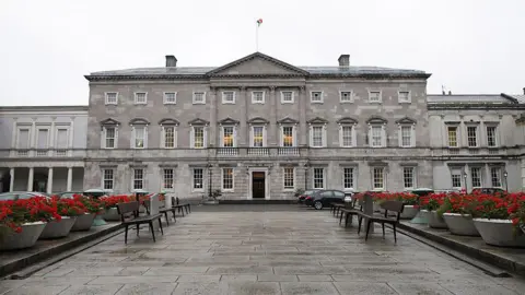 Getty Images A general view of Leinster House which houses the Seanad chamber, also known as the upper house of the Irish parliament, is pictured in Dublin, Ireland, in 2013. It is a large grey Georgian, Palladian style building with the Irish flag on top. A row of red flowers in pots and seating benches line a walkway leading up to it.