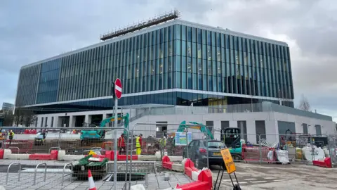 BBC The University of Bristol, Temple Quarter Enterprise Campus in Bristol. It is a very large modern-looking building with glass frontage and a University of Bristol sign on the top. In the foreground is construction work on the building. 