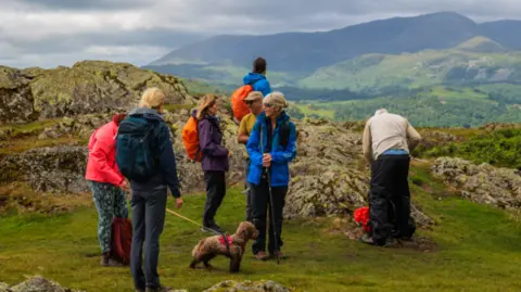 LDNPA A group of people are standing on the top of a crag with a landscape of mountains in front of them. One has a dog, one is standing apart looking, up two are chatting and one is looking into his backpack