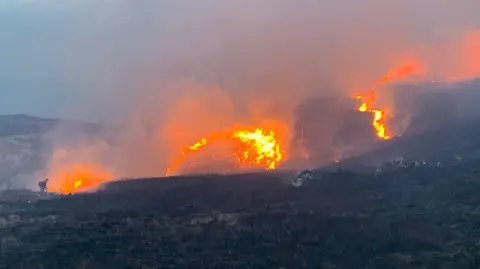 PA Media A gamekeeper from the Scottish Gamekeeper's Association uses a leaf blower to direct flames in a wildfire at the end of June - we see a dark hill with bright flames creeping up it and smoke filling the sky