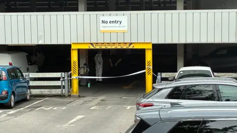A picture of the entrance to the car park. There is a yellow entrance with forensic teams pictured inside.
