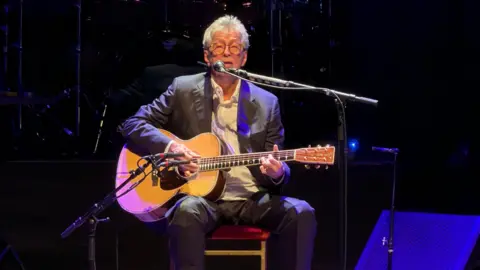 Lee Anderton A man, Eric Clapton, sat holding a guitar while performing on a stage.