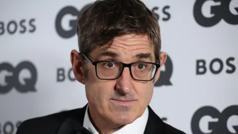 Lia Toby/Getty Images Louis Theroux is wearing a tuxedo. A backdrop is behind him. He is wearing black glasses. 