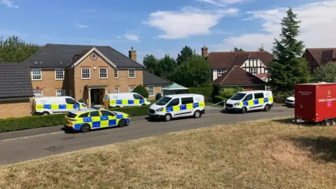 DJ McLaren/BBC About six police vehicles parked outside a large modern, two-storey brick house. Next door on the right is a mock Tudor house - both have separate garages in front of them. There is also a red vehicle/trailer with "Fire & Rescue Service" and "Welfare Support..." written on it.