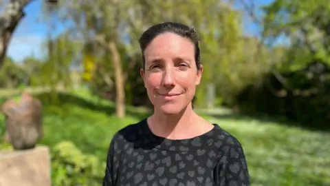 Woman in her 40s in a black jumper top with black hearts on it. She has brunette hair, back in a ponytail. she is stood in a luscious green garden