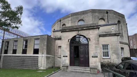 BBC An old court building with a big arched door, grey stone walls and lots of windows. Behind it is a modern building and a tree. 
