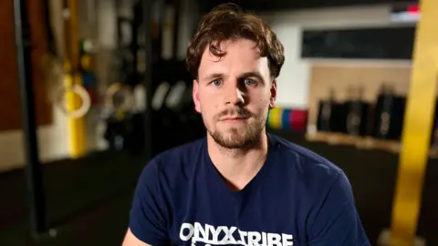 MARTIN GILES/BBC Louis Pearce looks at the camera in the Onyx Tribe Crossfit Gym in Huntingdon. You can see his head and shoulders and he is wearing a navy blue t-shirt. He has brown hair and a short beard. There are gym rings and bar bells in the blurred background.