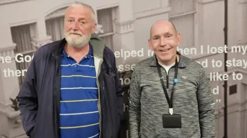Good Shepherd Joseph is stood with Paul with a charity poster as a backdrop. He has a blue striped T-shirt on and navy jacket and white beard. Paul has a grey top on with a lanyard on his chest. He has cropped hair.