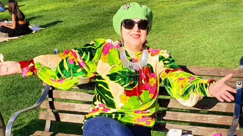 Handout A woman with a green hair covering, a bright green, red and yellow floral print blouse and jeans, wearing large sunglasses sits on a park bench in front of a grassy field smiling with her arms outstretched.