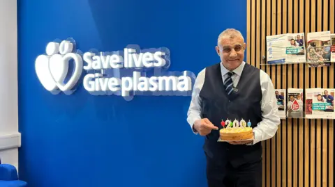 An older man who is wearing a shirt, waistcoat and suit trousers holding a cake. He is posing for a picture in front of a blue wall that had a white sign which reads "save lives give plasma". He is smiling.