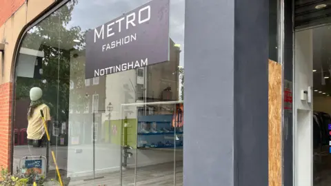 Shop frontage of Metro Fashion showing a large window and doorway, where part of the doorway glass has been boarded over