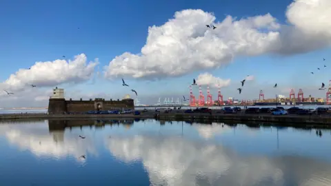 New Brighton's coastline photographed on a beautiful sunny day. The blue skies and white fluffy clouds are reflected in the calm water.