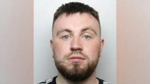 A police mugshot of Ethan Hallows. He has short, brown hair, closely shave at the side of his head. He has a dark, brown beard and facial hair.