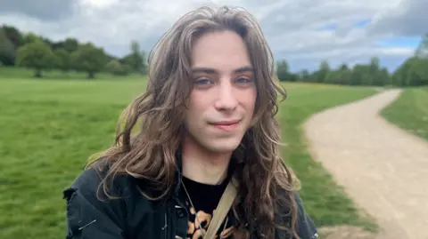Josh McLaughlin/BBC Fergus Chamberlain looks straight into the camera with a half-smile on his face. He has long wavy hair and is wearing a band t-shirt, jacket and a silver necklace rest below a crossbody bag. An open field lies behind him, with a dirt pathway trailing behind him. It is daytime. Deciduous trees line the horizon with dark clouds covering patches of a blue sky.