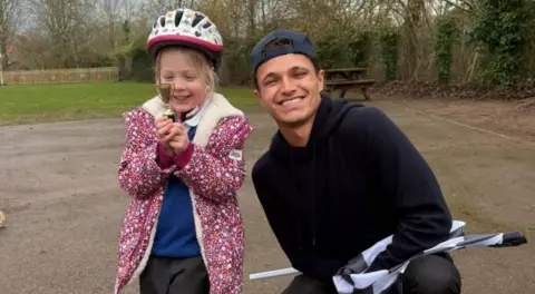 Lando Norris kneeling down and smiling broadly next to a schoolgirl wearing a multi-coloured pink coat and cycling helmet over her blue and grey uniform. She is looking at a miniature trophy and smiling. They are in a school playing area with a picnic bench in the background