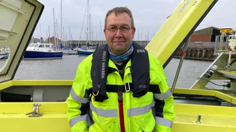 A man in a yellow high-vis jacket with a life vest stands with his hands behind his pockets in front of the yellow vessel. He has short brown hair and glasses.