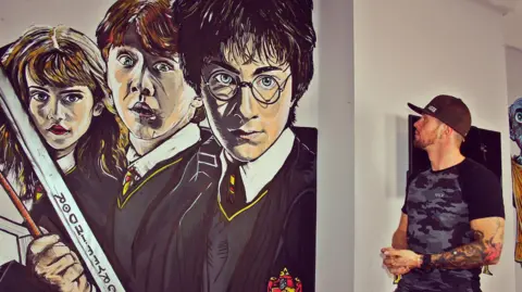 Adam Salisbury Adam Salisbury who has stubble wearing a black baseball cap and black and grey t-shirt stands by his Harry Potter mural on a bedroom wall.