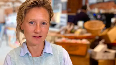 Martin Giles/BBC Charlotte Gurney is sitting down inside a farm shop at White House Farm in Sprowston. She is looking directly at the camera and smiling. She is wearing a lilac shirt with a grey sweater vest over the top.