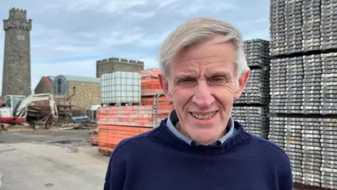 Smiling man in navy jumper, known as a "Guernsey Knit", stands in a construction yard with scaffolding, machinery, and stone tower behind him under an overcast sky.
